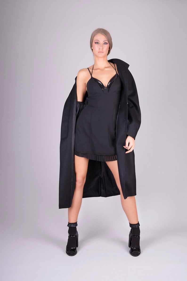 K&ouml;nnte beinhalten: Eine Frau in einem schwarzen Kleid und einem schwarzen Mantel. Sie steht vor einem grauen Hintergrund und blickt in die Kamera.