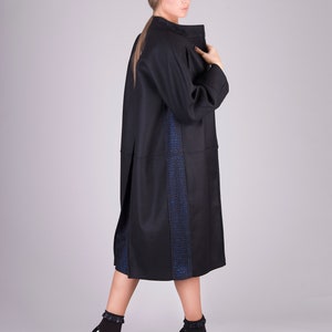 K&ouml;nnte beinhalten: Ein langer schwarzer Mantel mit einem blauen und schwarzen Muster an der Seite. Der Mantel wird von einer Person mit langen blonden Haaren getragen. Die Person tr&auml;gt schwarze Schuhe mit Absatz und R&uuml;schen.