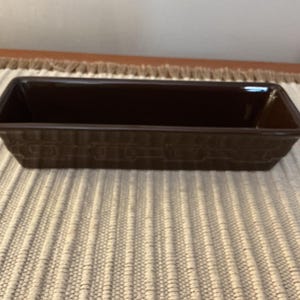 May include: A rectangular, dark brown ceramic loaf pan. The pan has a basket weave pattern on the sides and a smooth, glossy interior. The pan is empty and sits on a neutral-colored, textured surface.