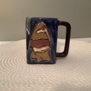 May include: A square ceramic mug with a dark blue background and a brown handle. The mug features a stylized design of a bird in shades of brown, tan, and maroon. The mug is sitting on a white, textured surface.