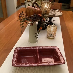 May include: A burgundy rectangular serving dish with two compartments sits on a white table runner. A decorative arrangement includes a pumpkin, candles, and fall foliage. The table is made of wood.