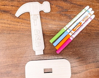 May include: A wooden hammer-shaped craft with the text "#1 DAD" and a rectangular base. Four colorful markers with the text "ALL ROADS LEAD TO ROME" are next to the hammer. The items are on a wooden surface.