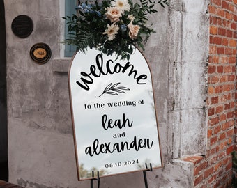 May include: A gold-framed arched mirror with a welcome sign for a wedding. The sign says "Welcome to the wedding of Leah and Alexander 08.10.2024".