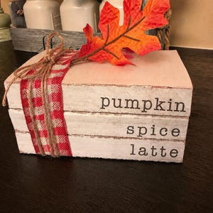 May include: A stack of three white wooden blocks resembling books, featuring the black printed text pumpkin, spice, and latte on the spines. The stack is tied with a red and white checkered ribbon and topped with a single orange and red faux leaf.