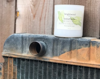 May include: White candle jar with a black and white label that reads "brightwood candle co. courage oak moss + amber hand poured soy candle 60 hour burn time burning for hope 9 oz"