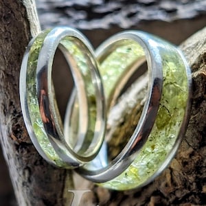 May include: Two silver rings with green gemstone inlays. The rings are stacked on top of each other and are sitting on a piece of wood. The text "Klaus Jewelers" is visible in the bottom right corner of the image.