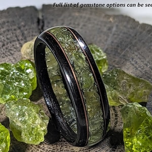 May include: A black ring with a green gemstone inlay and copper accents, size 8 1/2 US, width 6mm. The ring is surrounded by green gemstones. Text on the image says "Example Rings" and "Klaus Jewelers."