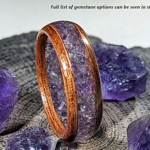 May include: A wooden ring with a band of amethyst gemstones set into the wood. The ring is 6mm wide and is shown on a wooden surface with amethyst stones.