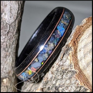 May include: A black wooden ring with a copper inlay and a center band filled with colorful opal fragments. The ring is set against a natural wood background, highlighting the contrast of materials and colors.