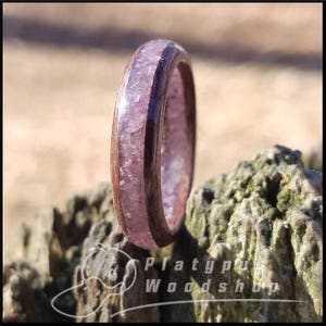 May include: A wooden ring with a band of dark brown wood and a center filled with a light purple, crystalline material. The ring is set against a blurred natural background. The ring is from Platypus Woodshop.