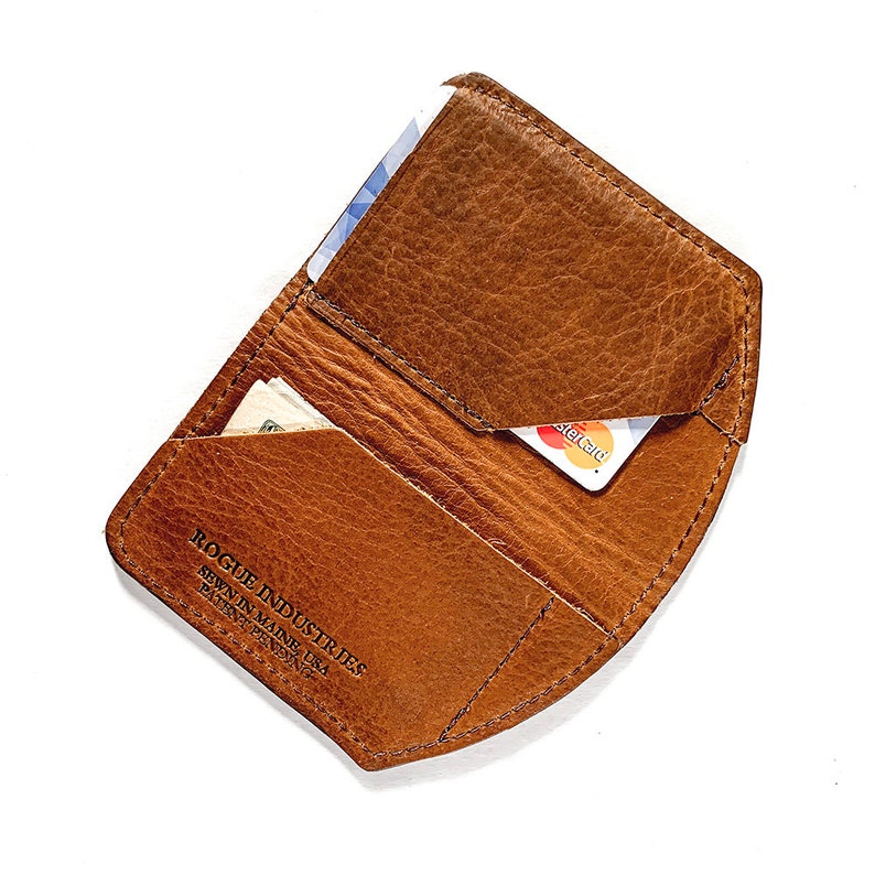 May include: Open brown leather wallet with card and cash slots. The wallet has a textured surface and visible stitching. The text "ROGUE INDUSTRIES SEBUN IN MAINE USA PATENT PENDING" is printed on the wallet.