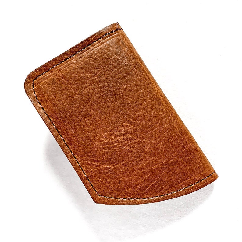 May include: A brown leather wallet with a textured surface. The wallet is rectangular with rounded corners and is stitched with black thread around the edges. The wallet is empty and is set against a white background.