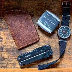 May include: An assortment of everyday carry items: a brown leather wallet, a black Leatherman multi-tool, a silver watch with a black strap, and a solid cologne. The cologne is labelled "FULTON & ROARK HATTERAS Classic, Greek, Spice."