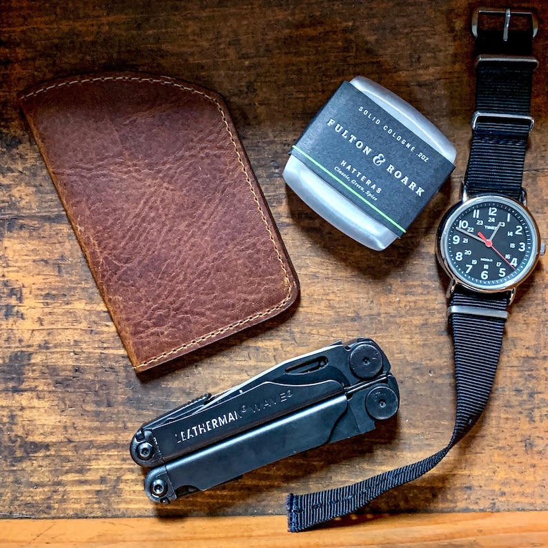 May include: An assortment of everyday carry items: a brown leather wallet, a black Leatherman multi-tool, a silver watch with a black strap, and a solid cologne. The cologne is labelled "FULTON & ROARK HATTERAS Classic, Greek, Spice."