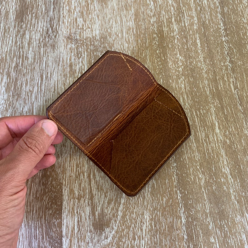 May include: A brown leather card holder with a textured surface and stitched edges. The open wallet is held in a hand, showcasing the interior pockets. The leather has a worn appearance.