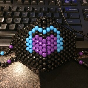 May include: A black beaded mask with a blue and purple heart design. The mask is made of small black beads and has a blue circle with a purple heart in the center.