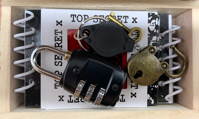 May include: A wooden box with a black and white notebook, a black combination lock, a brass padlock, and a black magnifying glass with a key ring. The notebook has the words "Top Secret" written on it.