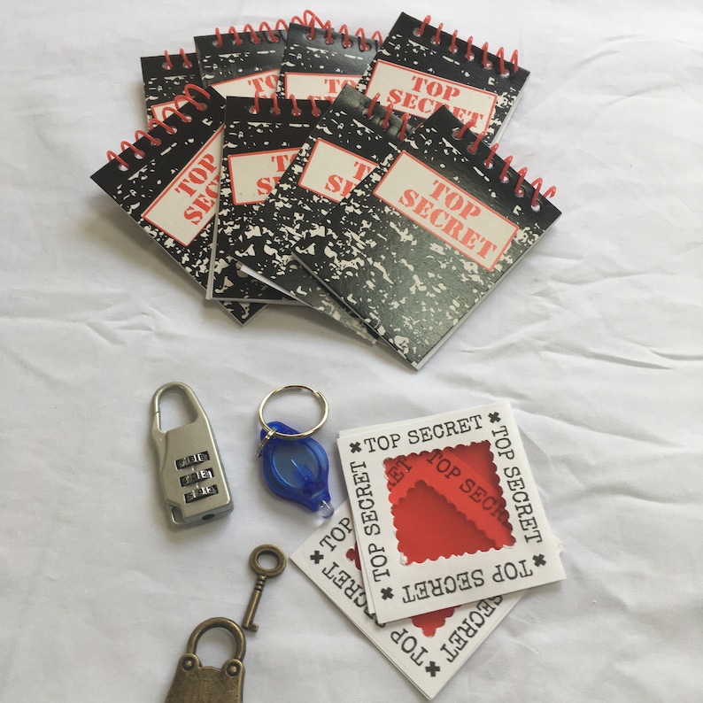 May include: A set of eight small black and white spiral notebooks with the words "Top Secret" printed on the front. The notebooks are on a white surface with a red and white "Top Secret" card, a blue key chain, a silver combination lock, and a gold key.