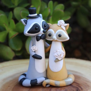 May include: A charming wedding cake topper featuring two raccoon figurines. The groom raccoon wears a top hat and bow tie, while the bride raccoon has a floral headpiece. Both hold roses, with a gray, white, and black color scheme.