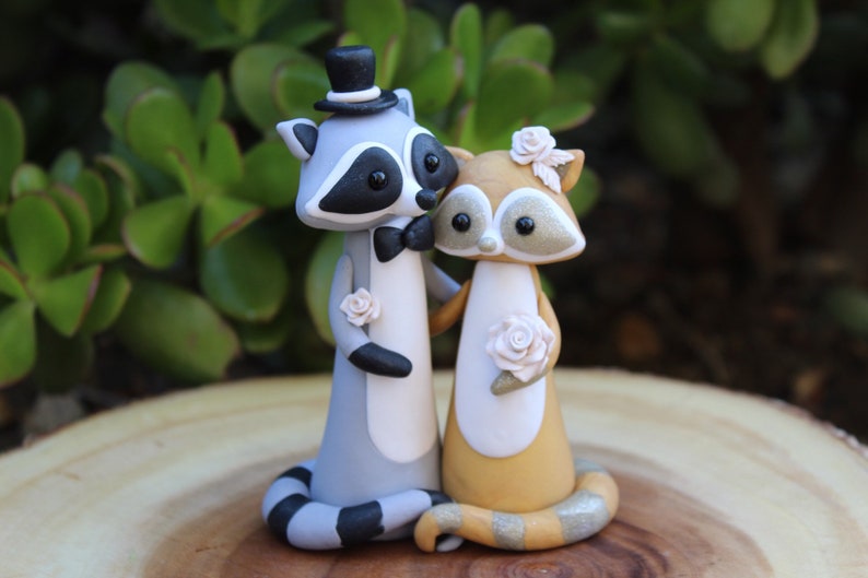 May include: A charming wedding cake topper featuring two raccoon figurines. The groom raccoon wears a top hat and bow tie, while the bride raccoon has a floral headpiece. Both hold roses, with a gray, white, and black color scheme.