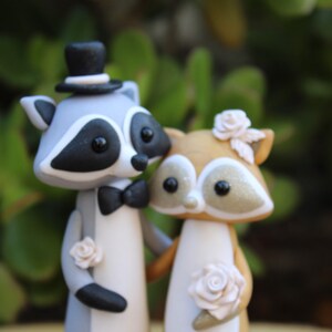 May include: A whimsical wedding cake topper featuring a raccoon groom in a top hat and bow tie, and a fox bride adorned with flowers. The figures are in shades of gray, brown, and white, with delicate floral accents.