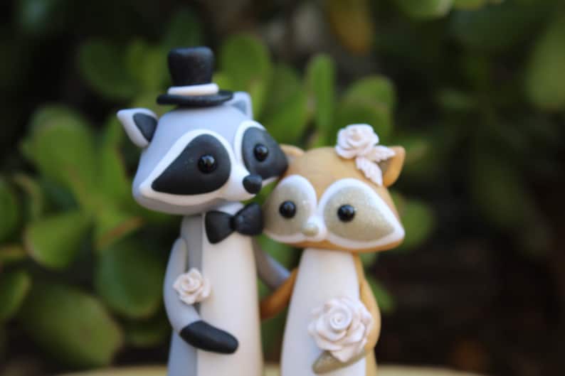 May include: A whimsical wedding cake topper featuring a raccoon groom in a top hat and bow tie, and a fox bride adorned with flowers. The figures are in shades of gray, brown, and white, with delicate floral accents.