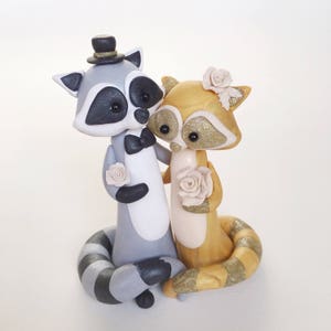 May include: A whimsical wedding cake topper featuring two raccoon figurines. One raccoon is gray with a top hat and bow tie, the other is gold with glitter accents. Both hold cream-colored roses, creating a romantic and celebratory theme.