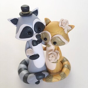 May include: A handcrafted wedding cake topper featuring two raccoon figurines. One raccoon is gray with a top hat and bow tie, the other is gold with glitter accents. Both raccoons hold cream-colored roses, perfect for a wedding.