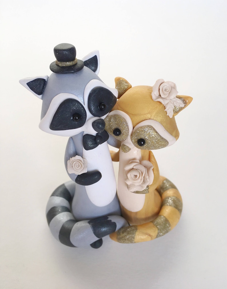 May include: A handcrafted wedding cake topper featuring two raccoon figurines. One raccoon is gray with a top hat and bow tie, the other is gold with glitter accents. Both raccoons hold cream-colored roses, perfect for a wedding.