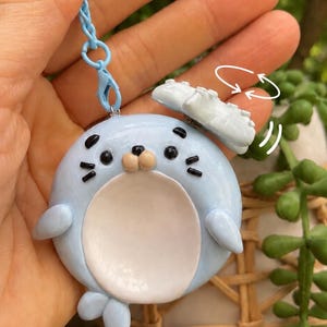 以下が含まれることがあります： 白いお腹と小さな白い雲が付いた、水色のあざらしのキーホルダー。アザラシは黒い目とひげがあります。キーホルダーは水色のチェーンで保持されています。