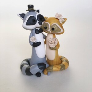 May include: A whimsical wedding cake topper featuring two raccoon figurines. The gray and white raccoon wears a top hat and bow tie, while the golden raccoon has a floral headpiece. Both hold roses, creating a charming, romantic scene.