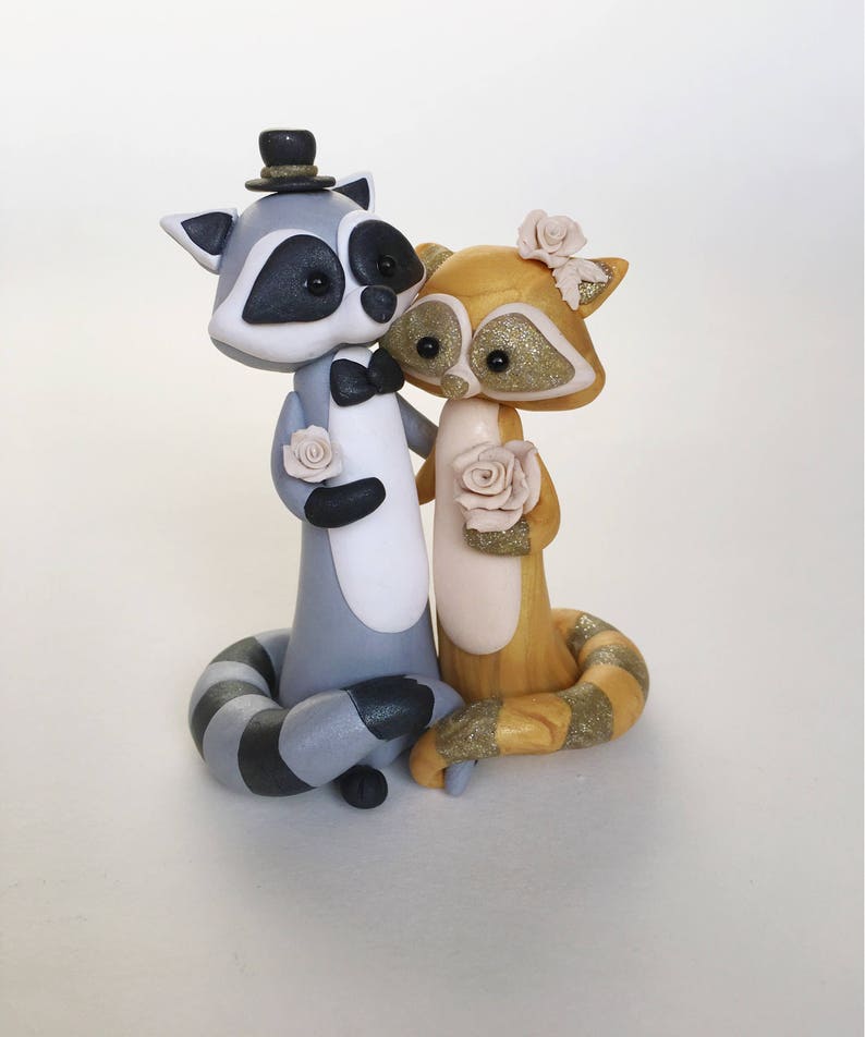 May include: A whimsical wedding cake topper featuring two raccoon figurines. The gray and white raccoon wears a top hat and bow tie, while the golden raccoon has a floral headpiece. Both hold roses, creating a charming, romantic scene.