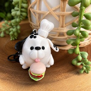 May include: A white dog figurine keychain wearing a chef hat and holding a small hamburger. The dog has black ears, black spots on its head, and black eyes. The hamburger features a pink bun, a brown patty, red tomato, and green lettuce layers.