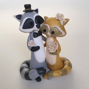 May include: A charming wedding cake topper featuring two raccoon figurines. One raccoon wears a top hat and bow tie, the other a flower in its hair. Both hold roses. The raccoons are gray, gold, and white with glitter accents.