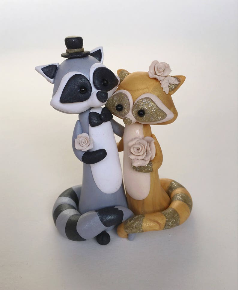 May include: A charming wedding cake topper featuring two raccoon figurines. One raccoon wears a top hat and bow tie, the other a flower in its hair. Both hold roses. The raccoons are gray, gold, and white with glitter accents.