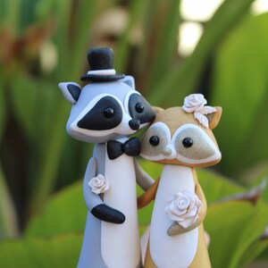 May include: A pair of raccoon figurines, one in a top hat and bow tie, the other with a floral headpiece and holding a rose. The figurines are gray, white, and tan, with black accents and glitter details. A unique wedding cake topper.