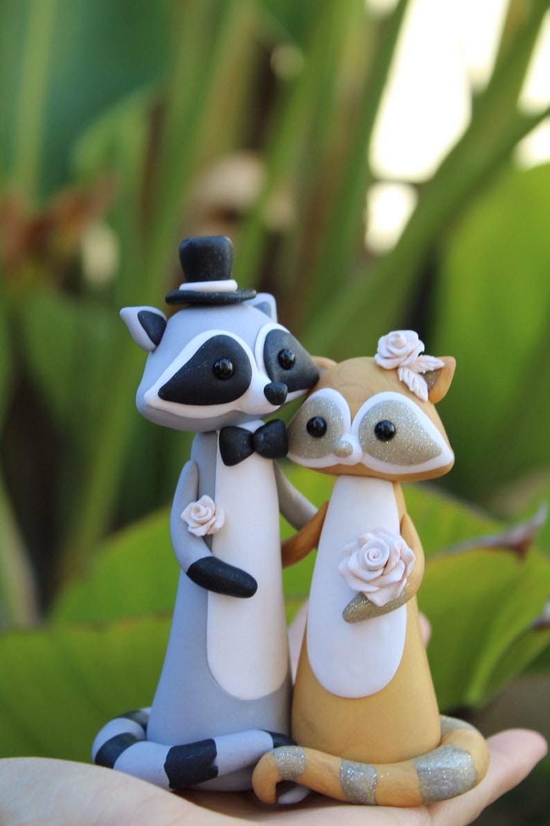 May include: A pair of raccoon figurines, one in a top hat and bow tie, the other with a floral headpiece and holding a rose. The figurines are gray, white, and tan, with black accents and glitter details. A unique wedding cake topper.