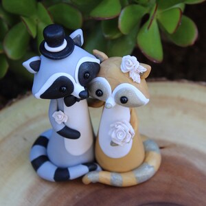 May include: A handcrafted wedding cake topper featuring two raccoon figurines. One raccoon wears a top hat and bow tie, the other has a floral headpiece. Both raccoons are holding each other, with floral accents.