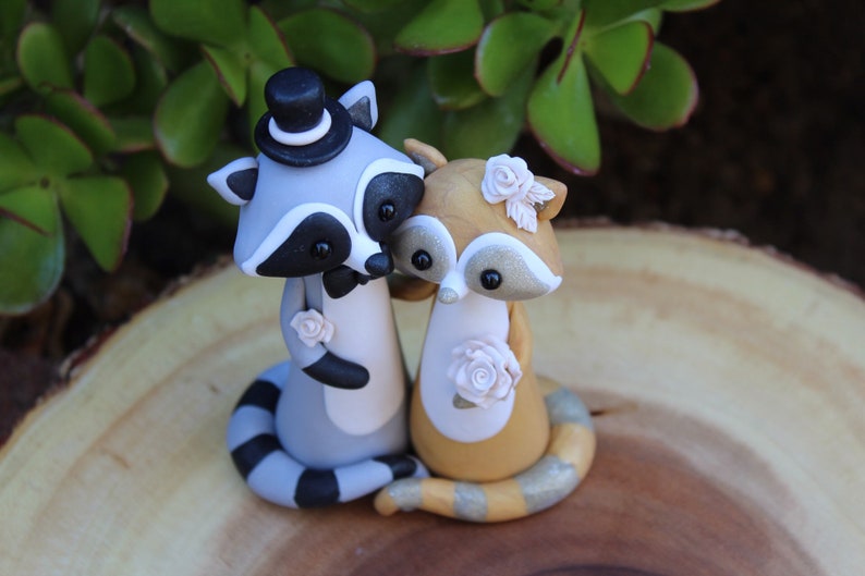 May include: A handcrafted wedding cake topper featuring two raccoon figurines. One raccoon wears a top hat and bow tie, the other has a floral headpiece. Both raccoons are holding each other, with floral accents.