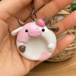 May include: A handmade, pink and white cow-shaped keychain. The cow has brown horns, a pink nose, and a strawberry on its head. The keychain is held by a person's hand. The cow is made of clay.