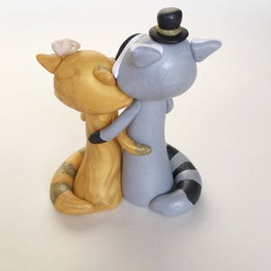 May include: A whimsical wedding cake topper featuring two raccoon figurines. One is gold with a flower, the other is gray with a top hat. They are embracing and kissing, with glitter accents on their arms and tails.