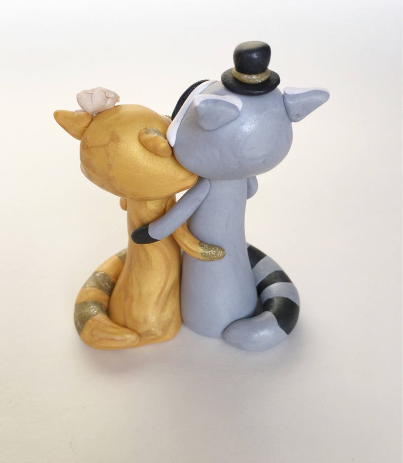 May include: A whimsical wedding cake topper featuring two raccoon figurines. One is gold with a flower, the other is gray with a top hat. They are embracing and kissing, with glitter accents on their arms and tails.