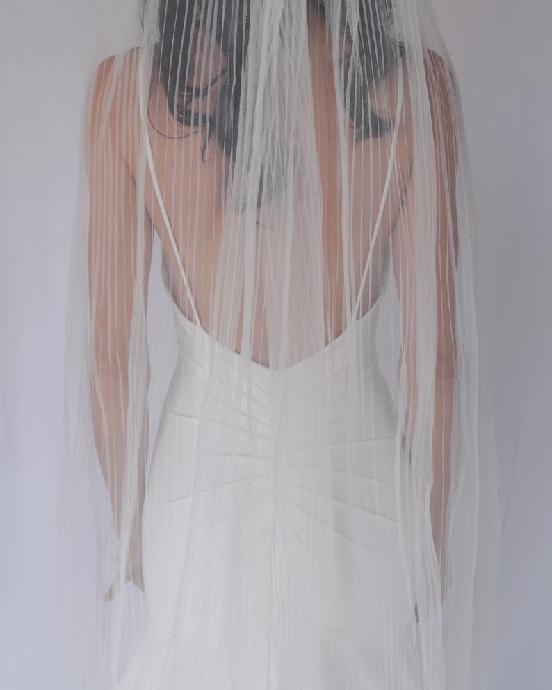 May include: A white wedding dress with a sheer veil. The dress has a low back and a fitted bodice.