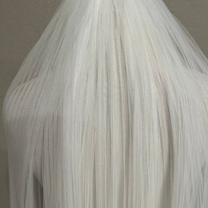 May include: A white tulle wedding veil with a single layer of soft, sheer fabric. The veil is gathered at the top and falls in a cascade of delicate pleats.