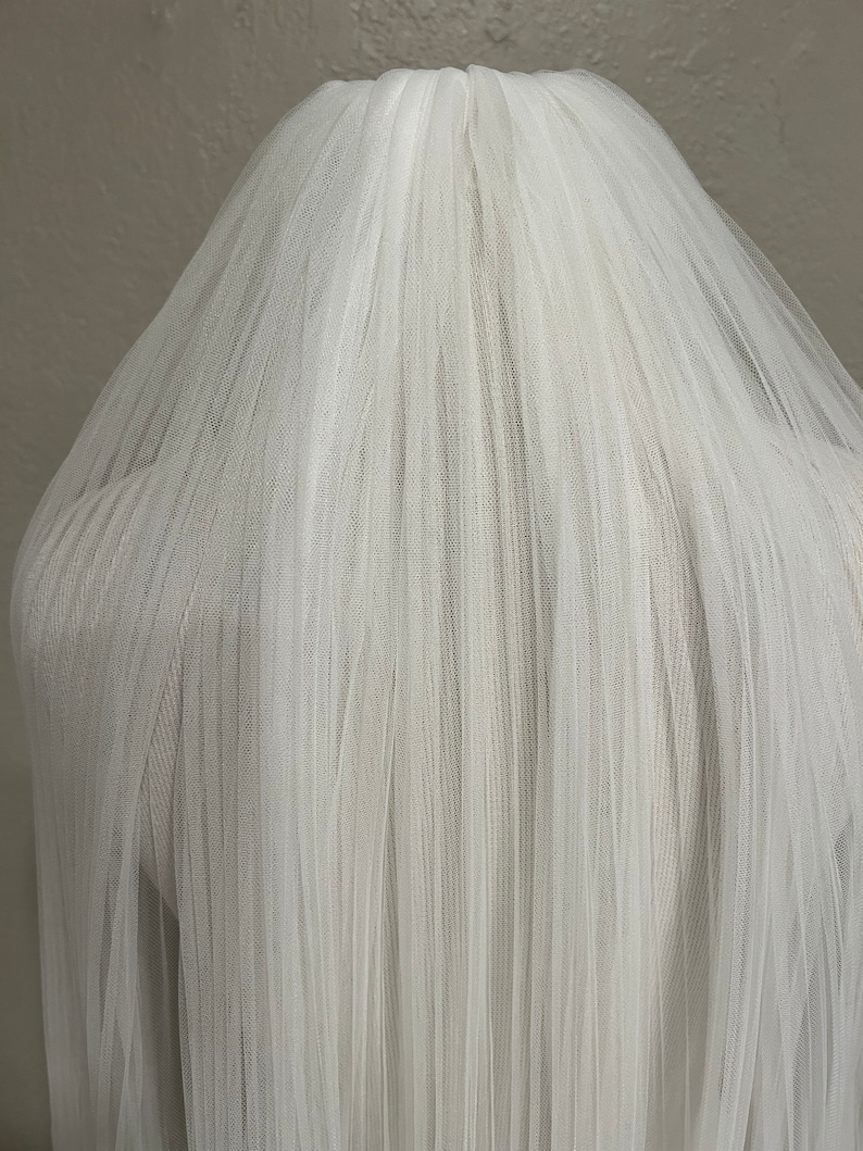 May include: A white tulle wedding veil with a single layer of soft, sheer fabric. The veil is gathered at the top and falls in a cascade of delicate pleats.
