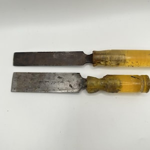 May include: Two metal wood chisels with flat, rectangular blades and translucent yellow plastic handles. The bottom chisel blade is stamped with the text FULLER'S TOOL STEEL USA. Both tools show signs of wear, including surface oxidation and discolouration.