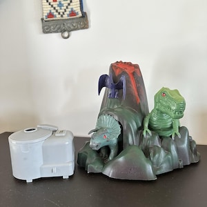 May include: A gray plastic projector device sits beside a molded plastic volcano playset. The volcano features a red lava peak, a green triceratops emerging from a cave, a green tyrannosaurus rex on the side, and a small purple pterodactyl.