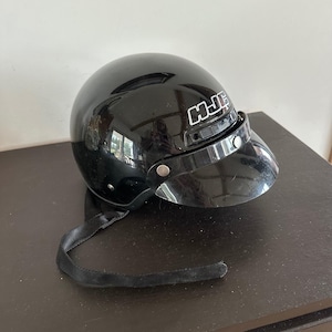 Op de afbeelding: Een glanzend zwarte HJC-motorhelm met een kort, donker getint vizier dat is bevestigd met zilverkleurige metalen drukknopen. De helm is voorzien van een zwarte kinband die aan de zijkant hangt en toont het witte HJC-logo aan de voorzijde.
