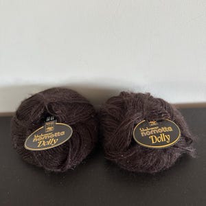 May include: Two round balls of dark brown yarn, each featuring an oval black label with gold text that reads Schachenmayr nomotta Dolly. The yarn has a fuzzy, textured appearance, and the labels are securely tucked into the center of each ball.