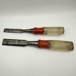 May include: Two wood chisels with weathered metal blades and red painted ferrules. The handles are made of clear, textured plastic, showing signs of age and use. The metal surfaces exhibit oxidation and surface wear consistent with manual tools.